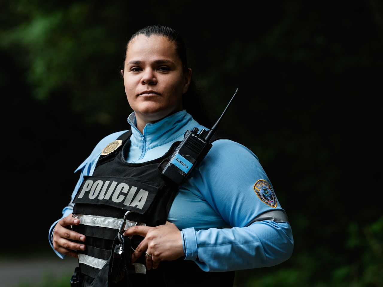 services-02 Female police officer in uniform standing outdoors with a confident expression and radio handset.